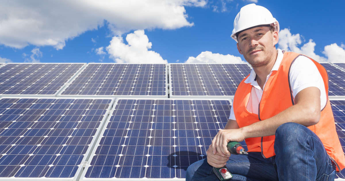 A Clean Energy Council Accredited Installer sits on a roofnext to solar panels with a drill in his hand, wearing a fluro vest and a white hard hat.